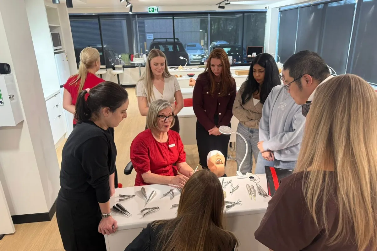Instructor in red demonstrates dental instruments to students around a clinic table with a dental mannequin head on display in a bright classroom period environment.