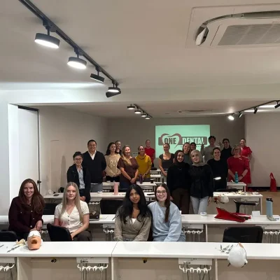 Group of people in a dental training classroom posing for a photo; dental mannequins and equipment visible on desks in the foreground.