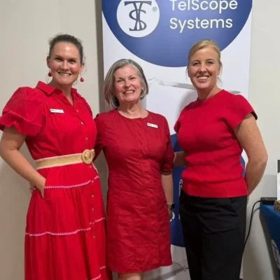 Three women in red outfits smile at the camera, posing in front of a blue and white banner that reads 'Telescope Systems'.