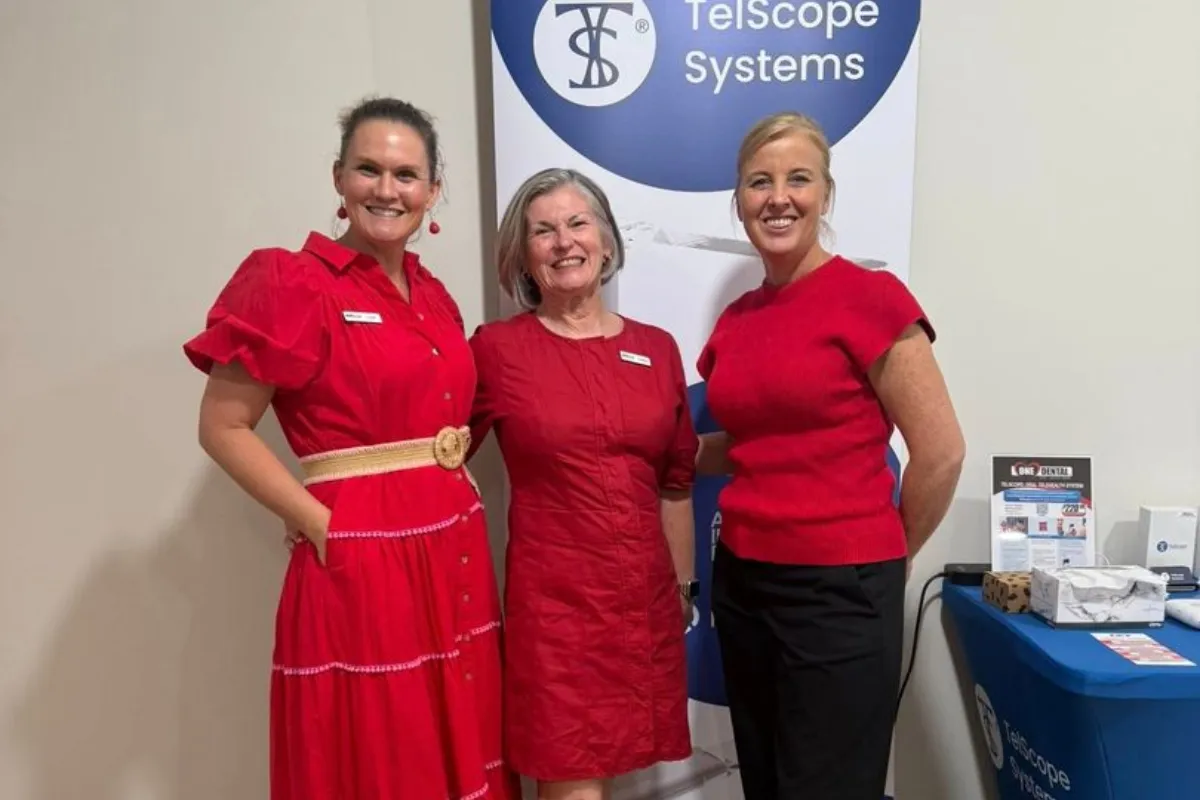Three women in red outfits smile at the camera, posing in front of a blue and white banner that reads 'Telescope Systems'.