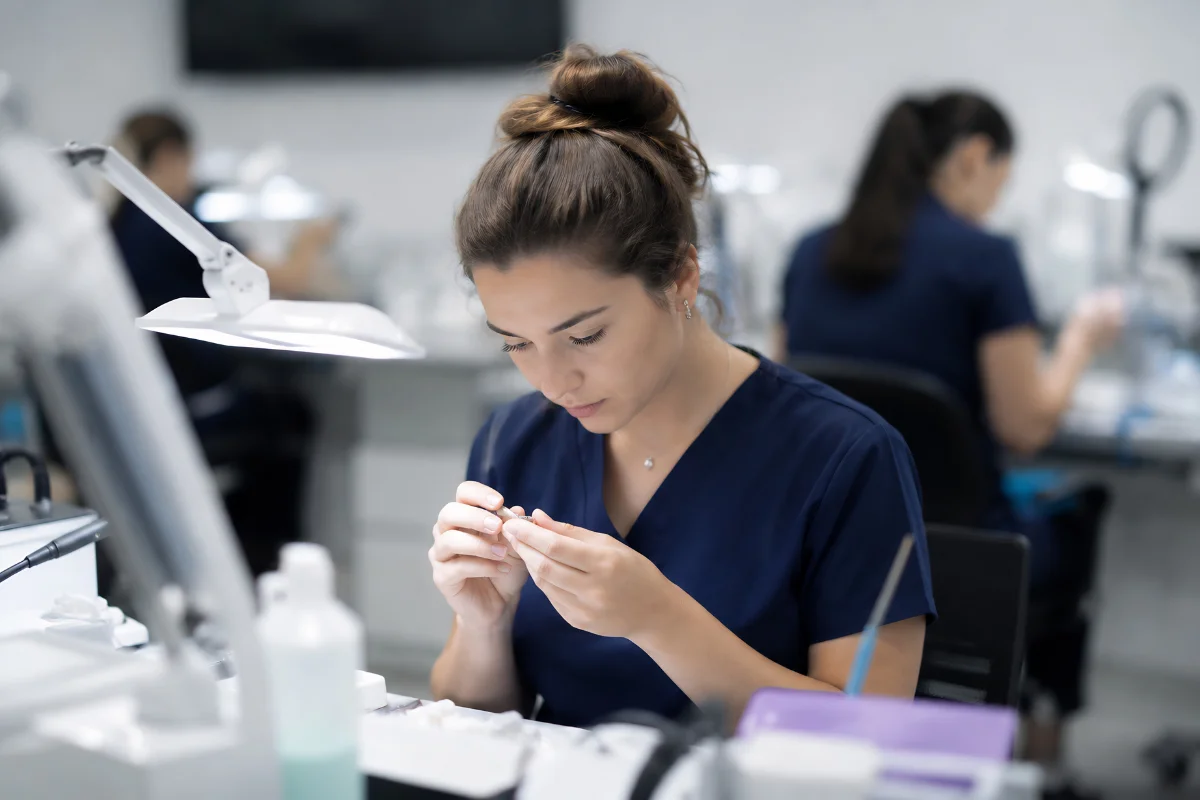 Female dental technician examining a dental model in a modern laboratory while colleagues work in the background.