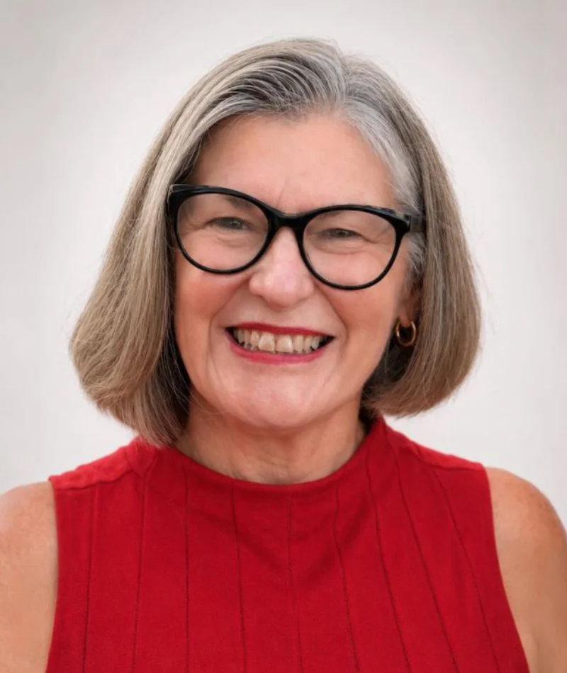 Smiling older woman with gray hair and black glasses wearing a red sleeveless top, gold hoop earrings, neutral background.