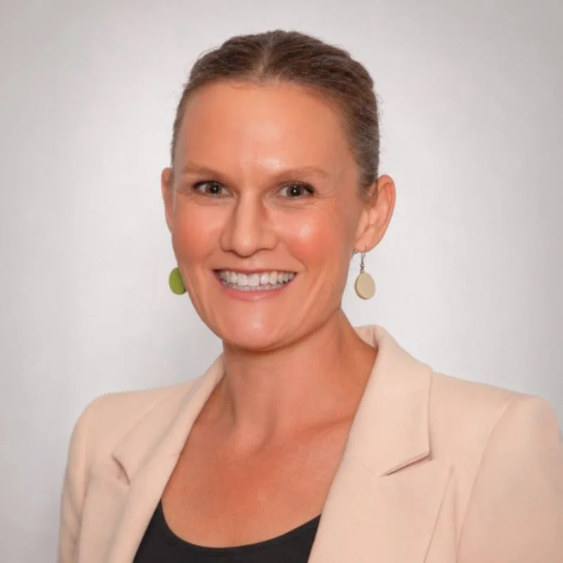 Head-and-shoulders portrait of a smiling woman in a beige blazer and black top, wearing oval earrings with a light background.