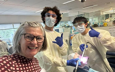 Smiling woman with glasses in a clinic pose with two staff in full PPE standing behind her, each giving a thumbs up; one holds a medical instrument.