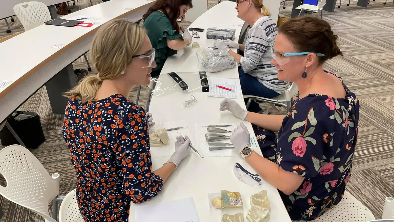 Dental students at a table handling dental instruments with gloves and safety glasses on a classroom setup.